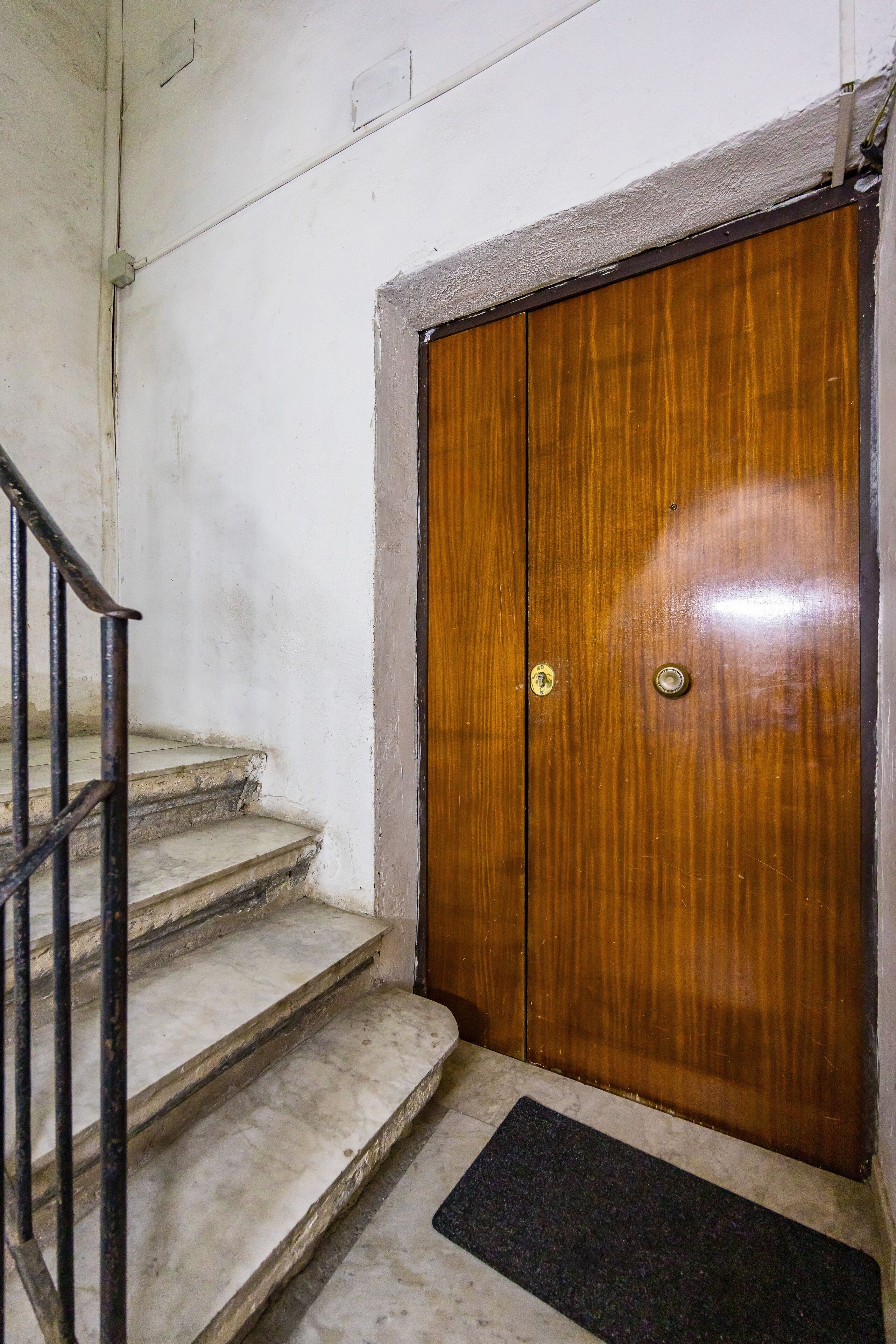 A wooden door with a metal handle, a black doormat, and a marble staircase with a metal railing.