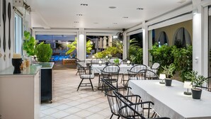 En indendørs patio med borde og stole, en pool i baggrunden og krukker med planter.