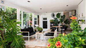 En rummelig opholdsstue med sofa, sofabord og stole, omgivet af store vinduer og indeplanter.