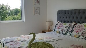 A bedroom with a floral bedspread, a swan-shaped pillow, a grey tufted headboard, and a window with a view of trees. - Kasa and family 220 (Valdivia)