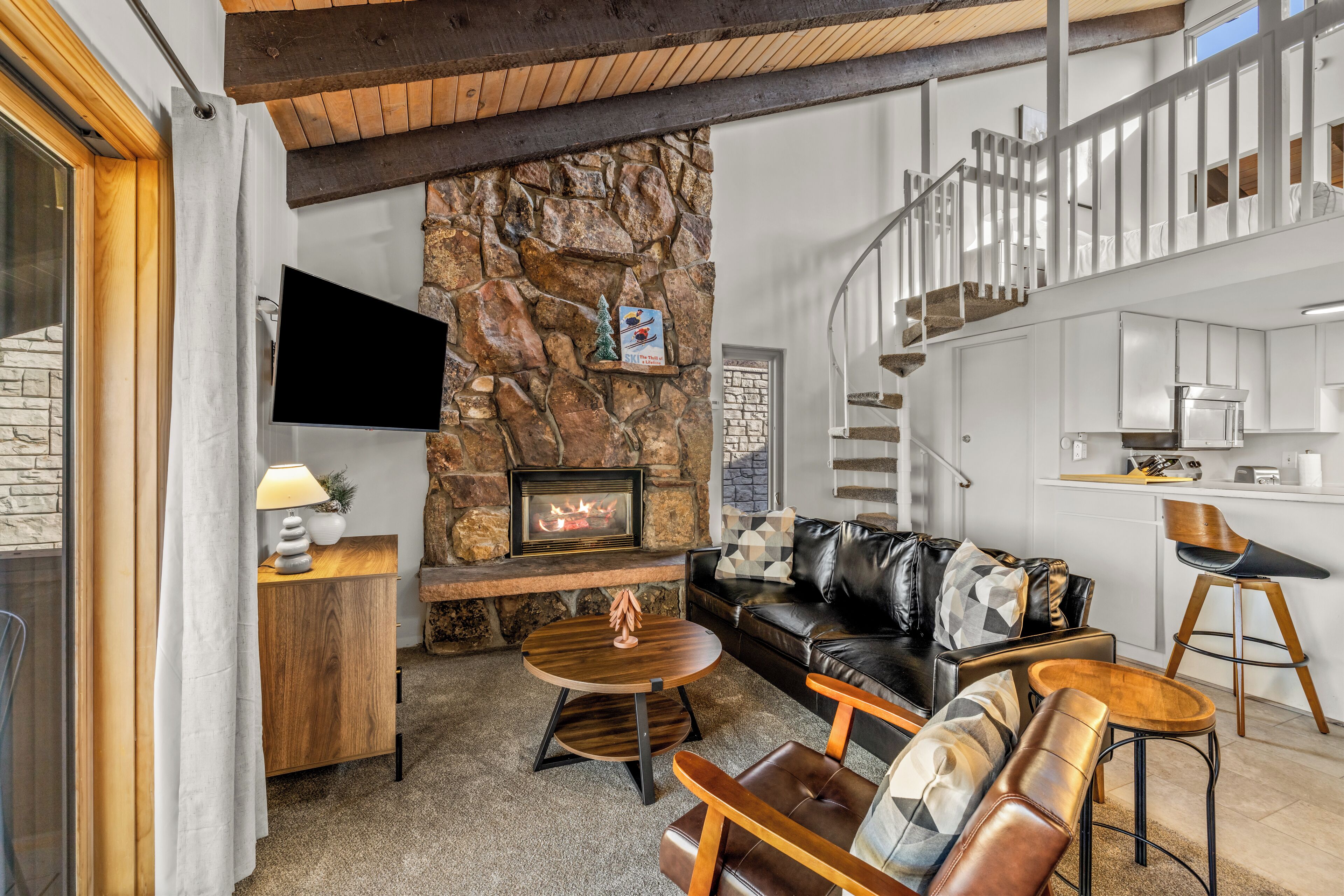 A modern living room with a stone fireplace, leather furniture, and a kitchen area with white cabinets.