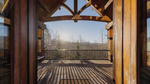 Una terraza de madera con barandilla de metal, vista a árboles y montañas al fondo, y un cielo despejado.