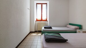 Une petite pièce propre, avec deux lits, une fenêtre avec des rideaux, un radiateur et un mur blanc.
