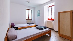 Une chambre de dortoir avec deux lits simples, un radiateur, une fenêtre avec des rideaux, une armoire en bois et un luminaire au plafond.