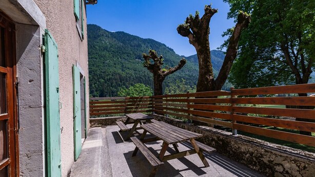 Un banc en bois, des tables de pique-nique, un mur de pierre et une vue sur une montagne boisée.