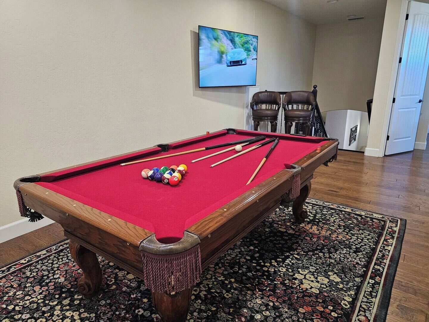 Een pooltafel met een rode viltkleed, omringd door een getextureerd vloerkleed, in een kamer met houten vloeren en een aan de muur bevestigde televisie.