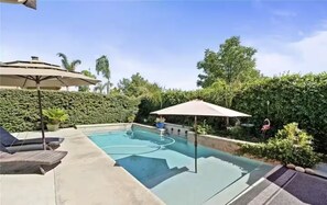 A swimming pool with lounge chairs and umbrellas, surrounded by greenery and a clear sky. - Urban comfort: Home w/ Game Room & Backyard Oasis (Clovis)