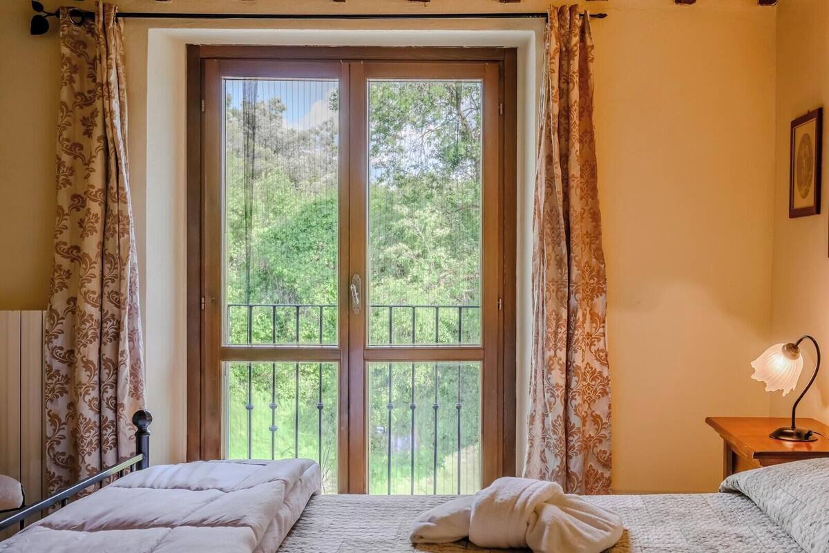 A bedroom with a large window, a bed with a white duvet, a bedside table with a lamp, and a framed picture on the wall.