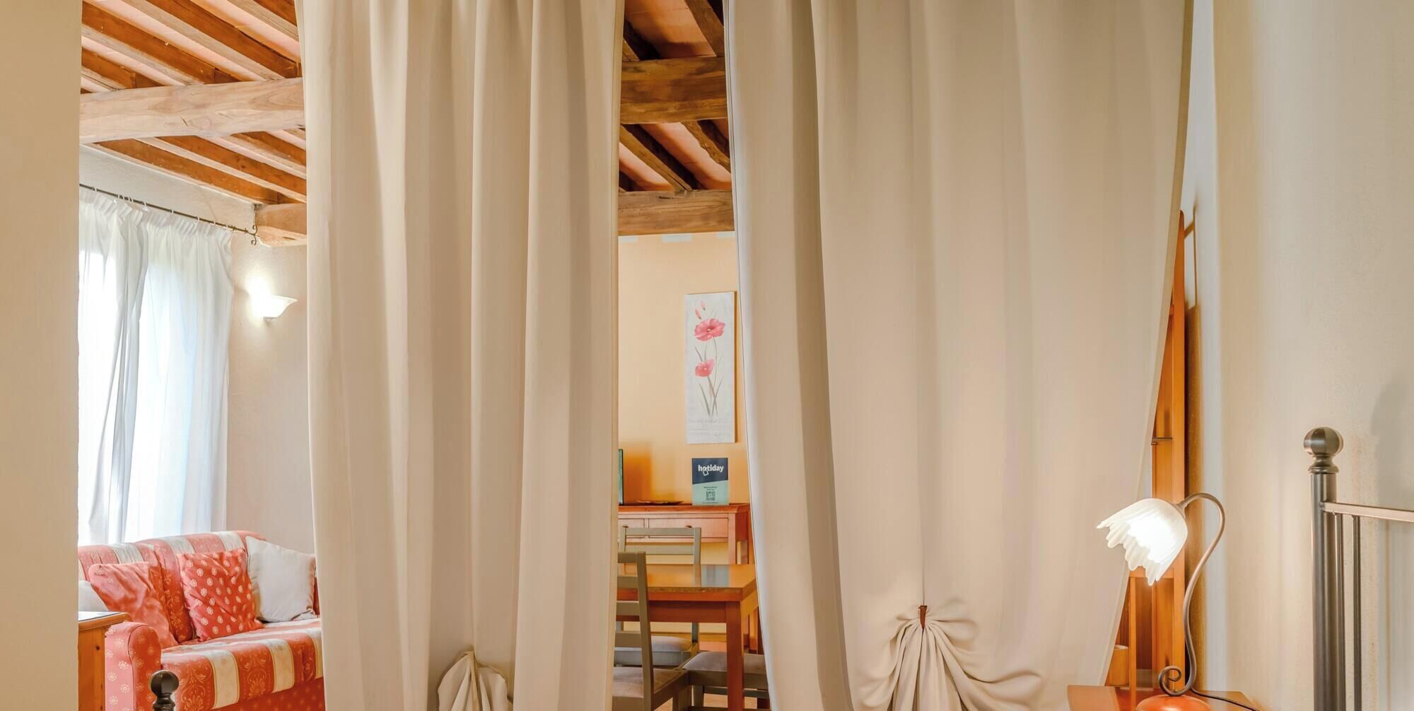 A room with a bed, a desk, and a chair. There is a window with curtains, a lamp, and a framed picture on the wall.