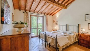 Ein Schlafzimmer mit Bett, Holz-Kopfstütze, Nachttisch, Fenster mit Vorhängen, Tür und Blick ins Grüne.