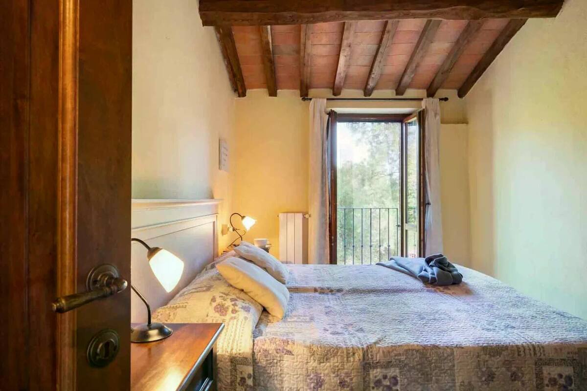 A bedroom with a wooden ceiling, a bed with a patterned bedspread, two bedside lamps, and a window with a view of trees.