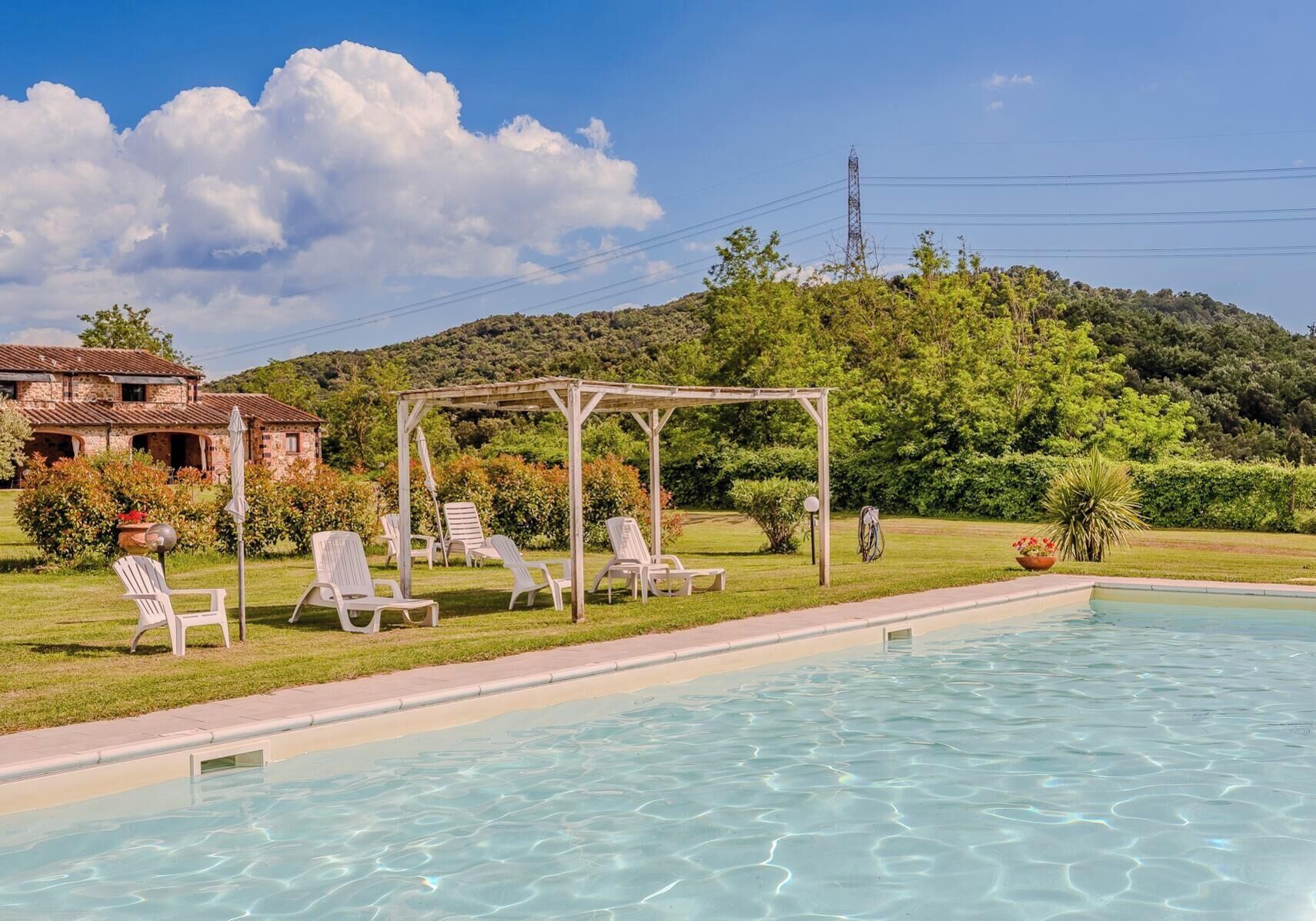 Ein Swimmingpool mit Liegestühlen und einer Pergola, umgeben von Grünpflanzen und einer Hügelkulisse.
