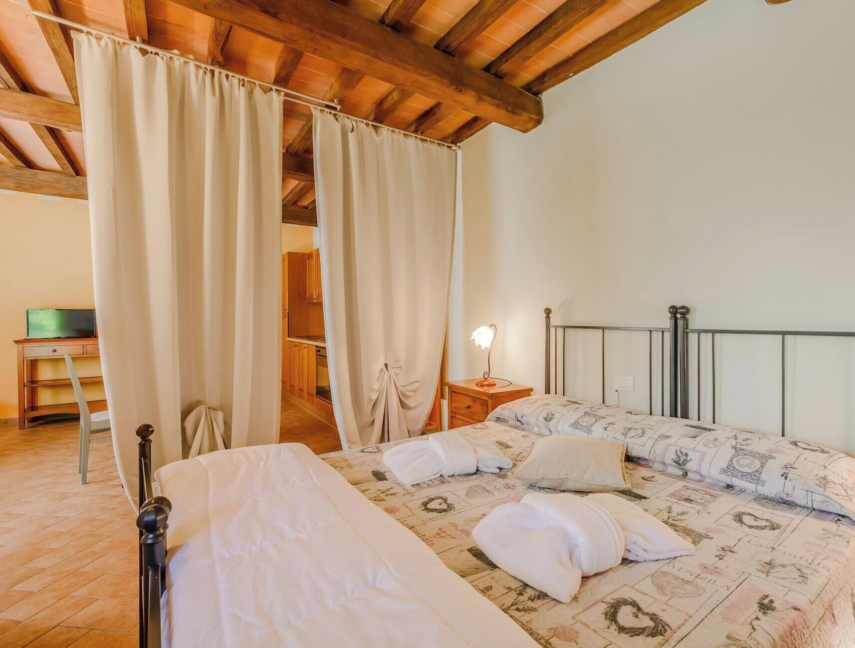 A bedroom with a bed, a nightstand, a lamp, and a view of a kitchen through a curtain.
