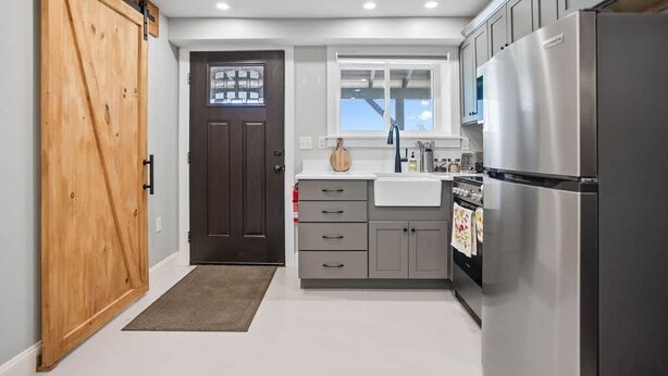 A compact kitchen with a wooden door, grey cabinets, a stainless steel refrigerator, and a window above the sink. - Raven Feather Retreat : Unit 2 (Liberty Township)