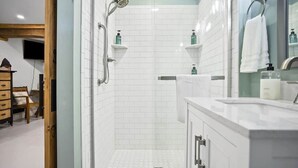 A modern bathroom with white tiles, a glass shower enclosure, a vanity with a mirror, and a towel rack. - Raven Feather Retreat : Unit 2 (Liberty Township)
