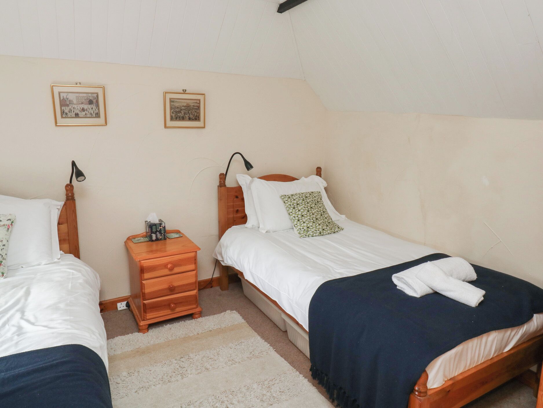 Un dormitorio con dos camas individuales, una mesita de noche con lámpara y dos cuadros enmarcados en la pared.