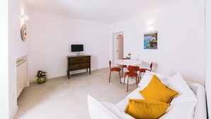 Una stanza con un divano bianco, un tavolino con sedie, una TV su un mobile in legno e un quadro appeso al muro.
