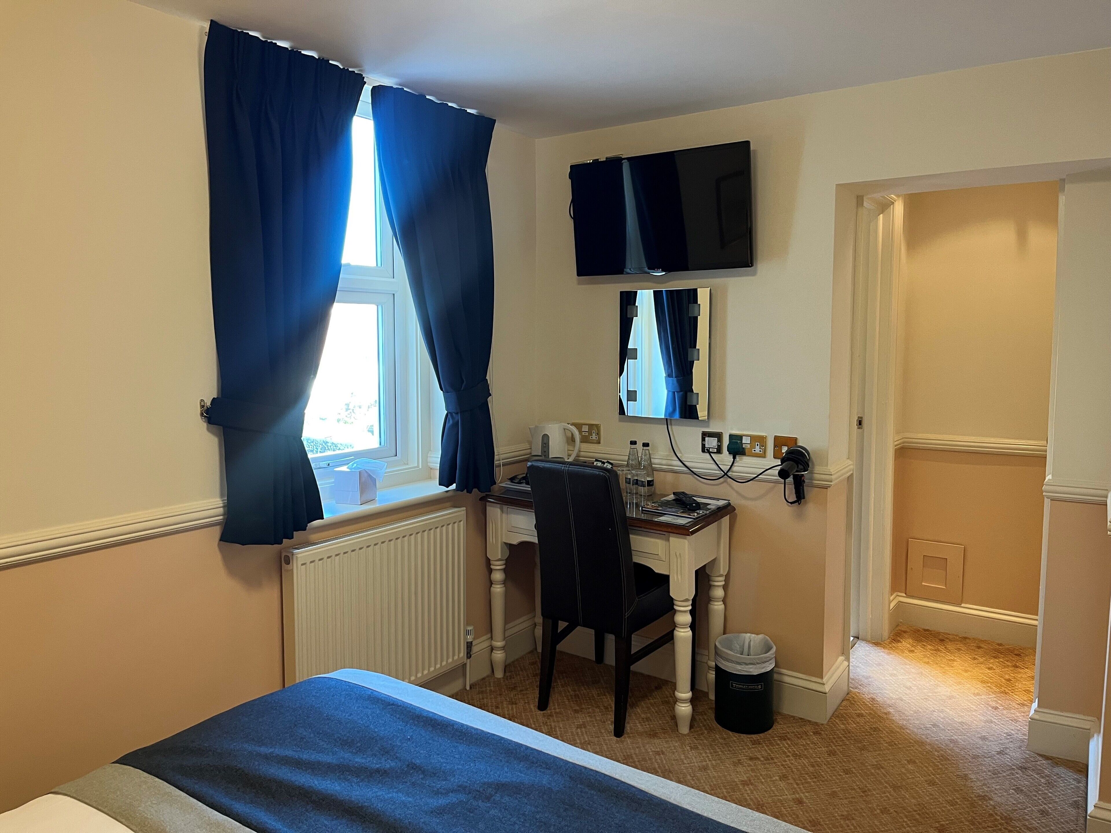 Una habitación de hotel con cama, escritorio con silla, televisor montado en la pared y ventana con cortinas.