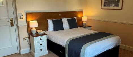 A hotel room with a large bed, two bedside tables with lamps, a nightstand, a drawer unit, and a framed picture on the wall.