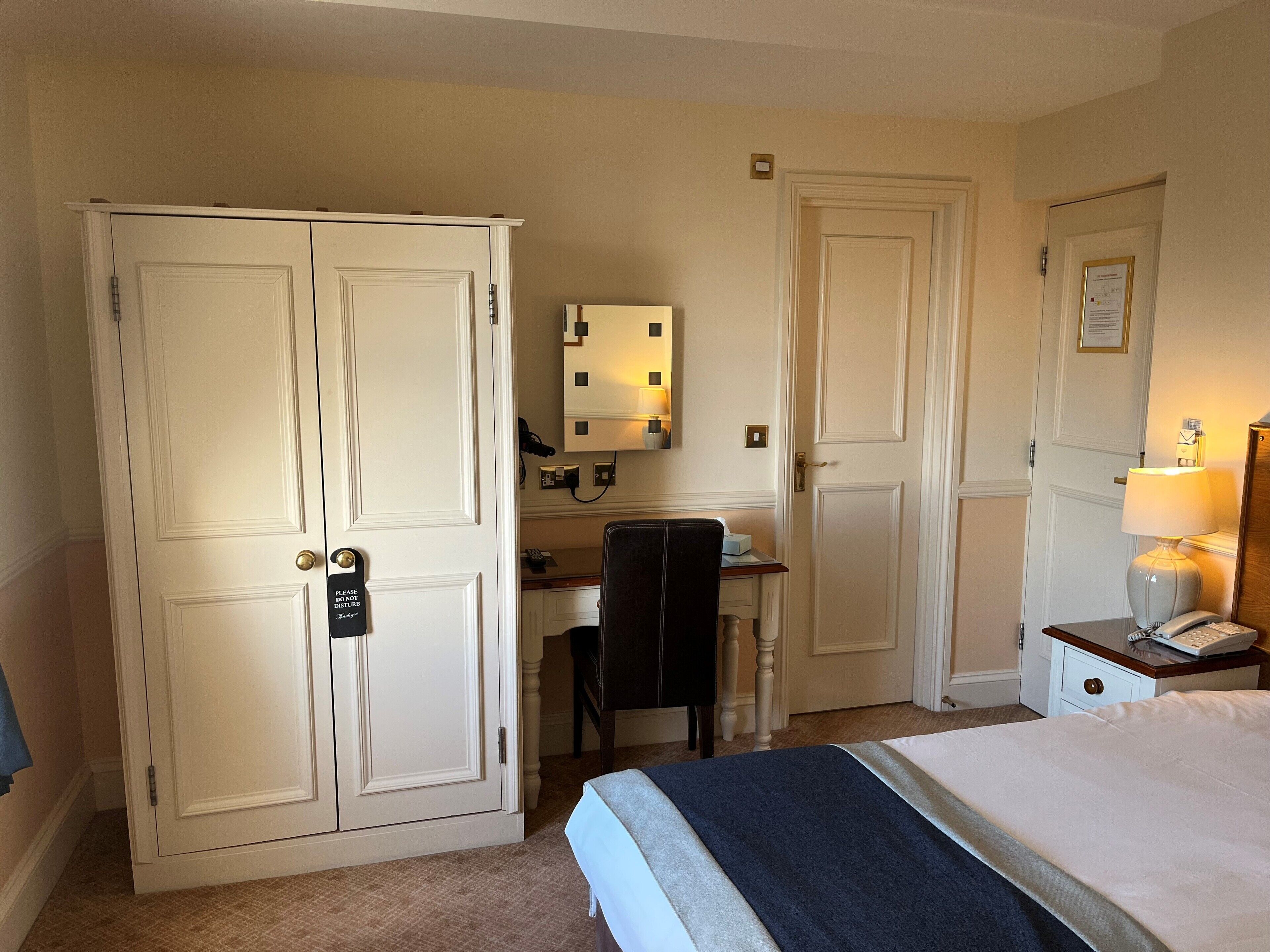 Una habitación de hotel con una cama, un escritorio con silla, un armario, una mesita de noche con lámpara y un teléfono.