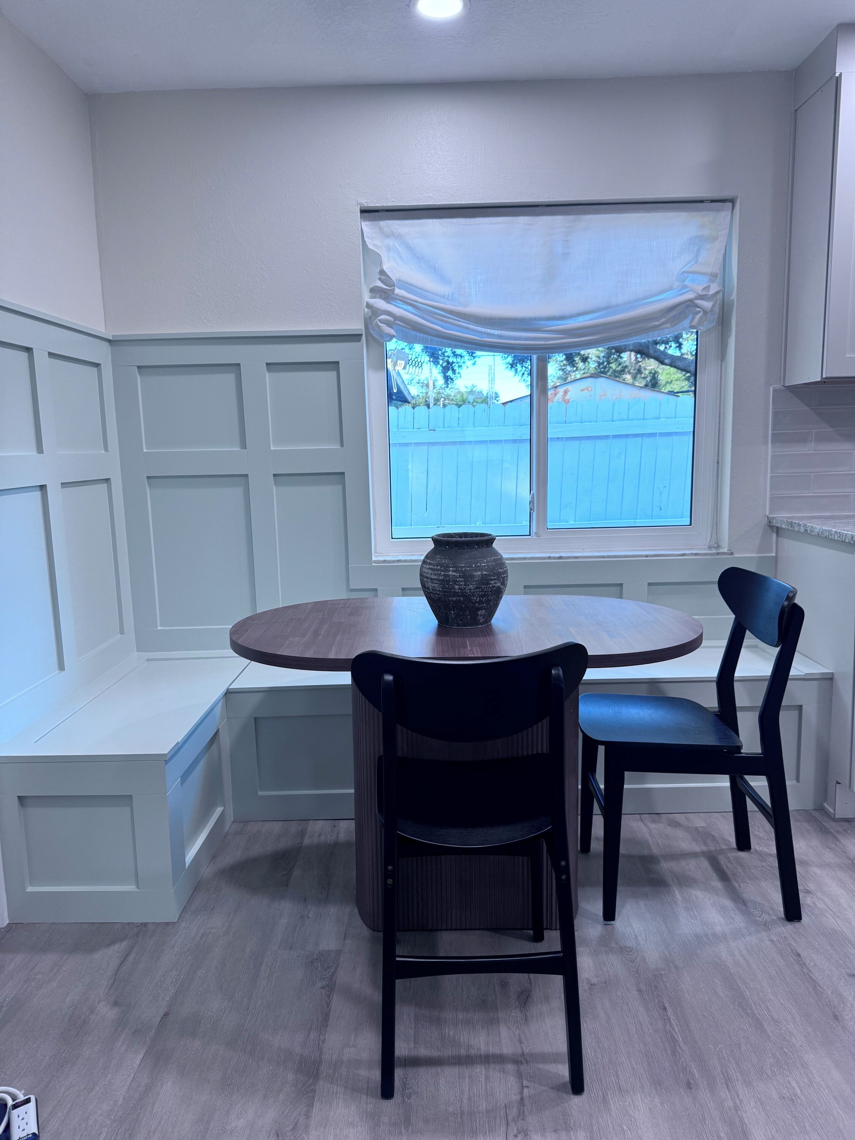 Una cucina con un tavolo rotondo, due sedie, una finestra con vista su una recinzione blu e un vaso sul tavolo.