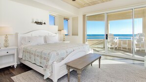 A bedroom with a large bed, a nightstand, a bench, and a view of the ocean through a sliding glass door.