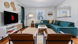 A modern living room with a flat-screen TV, a green sofa, a coffee table, and two brown chairs. - Chic 3BR Retreat•Firepit•Minutes to NC State (Raleigh)