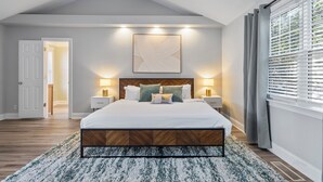 A bedroom with a large bed, two bedside tables with lamps, a ceiling light, a window with blinds, and a door leading to another room. - Chic 3BR Retreat•Firepit•Minutes to NC State (Raleigh)