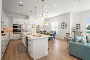 Una cocina moderna con armarios blancos, una isla central, iluminación colgante y un rincón de comedor con un sofá azul y una mesa.