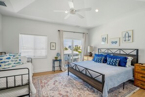 A bedroom with a bed, a bedside table, a nightstand, a window with blinds, a ceiling fan, and a view of the outside.