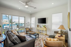 A spacious living room with a sofa, coffee table, and TV. There is a balcony with a view of houses.