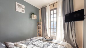 Een slaapkamer met een bed, een televisie, een raam met gordijnen, een plank met planten en uitzicht op het omliggende landschap.