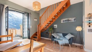 A room with a wooden staircase, a sofa, a dining table, and a view of mountains through an open door. - Gîte le Tech Nordic Bath & Sauna in the Heart of the Mountains (Arrens-Marsous)