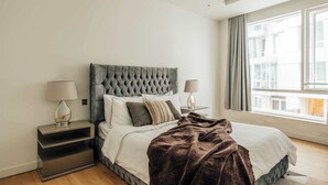 Ein modernes Schlafzimmer mit einer gesteppten Kopfteil, einem großen Fenster mit Vorhängen, einem Nachttisch mit Lampe und einem Bett mit brauner Decke.
