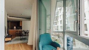 Ein modernes Wohnzimmer mit einem türkisfarbenen Sessel, einem Glastisch und Blick auf eine Stadtlandschaft durch große Fenster.