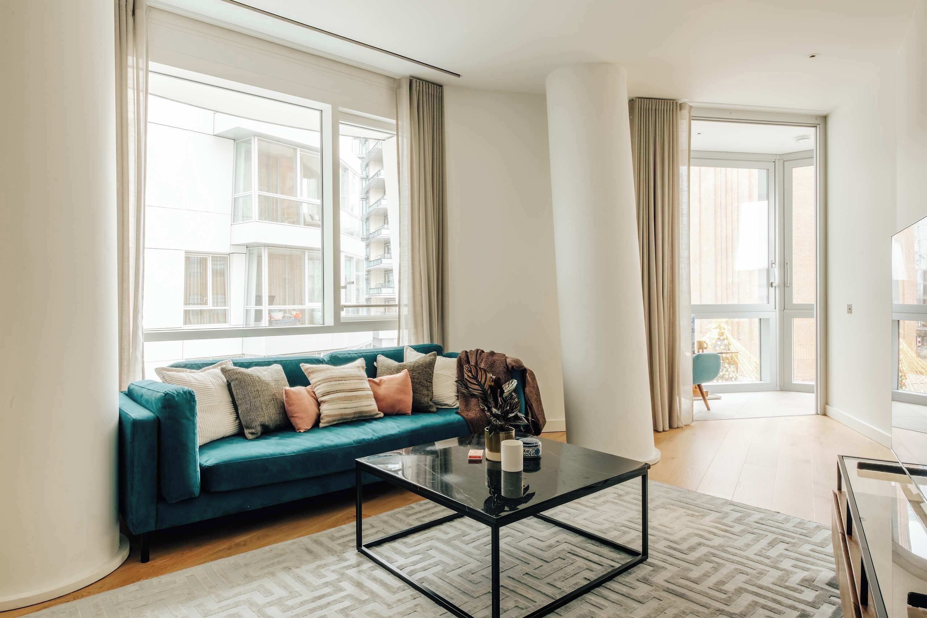 Un salon moderne avec un canapé de couleur turquoise, une table basse en verre et une vue sur un immeuble à travers de grandes fenêtres.