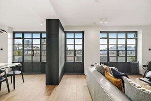 A modern living room with a sofa, a dining table with chairs, and large windows offering a city view. - Kensington Duplex w/ Private Terrace & portable AC (Greater London)