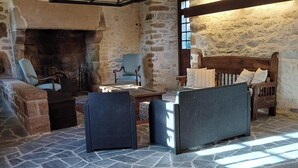 Una habitación con suelo de piedra, chimenea, muebles de madera y techo alto con vigas a la vista.