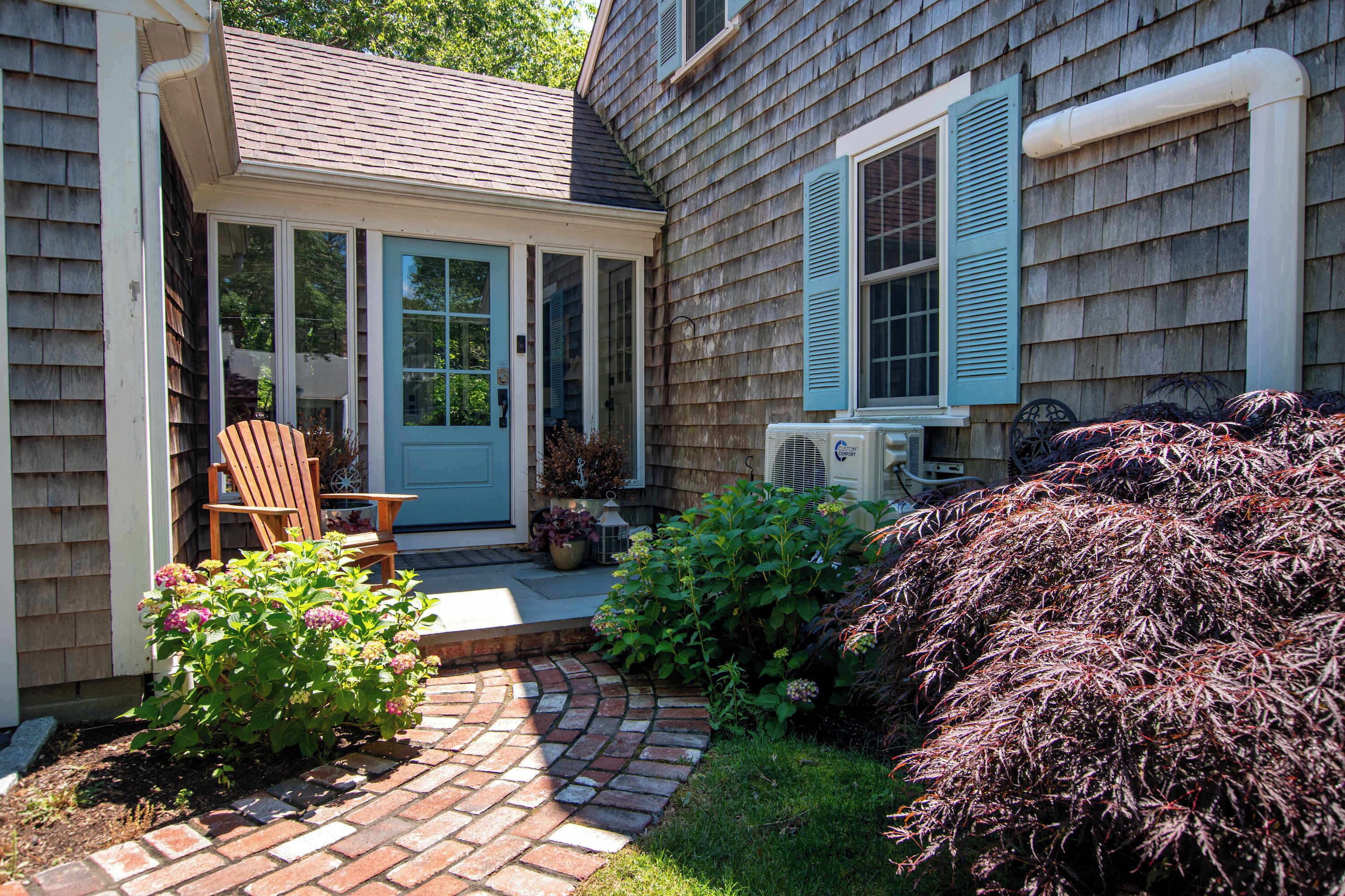 A house with a brick patio, a covered porch, a chair, and various plants.