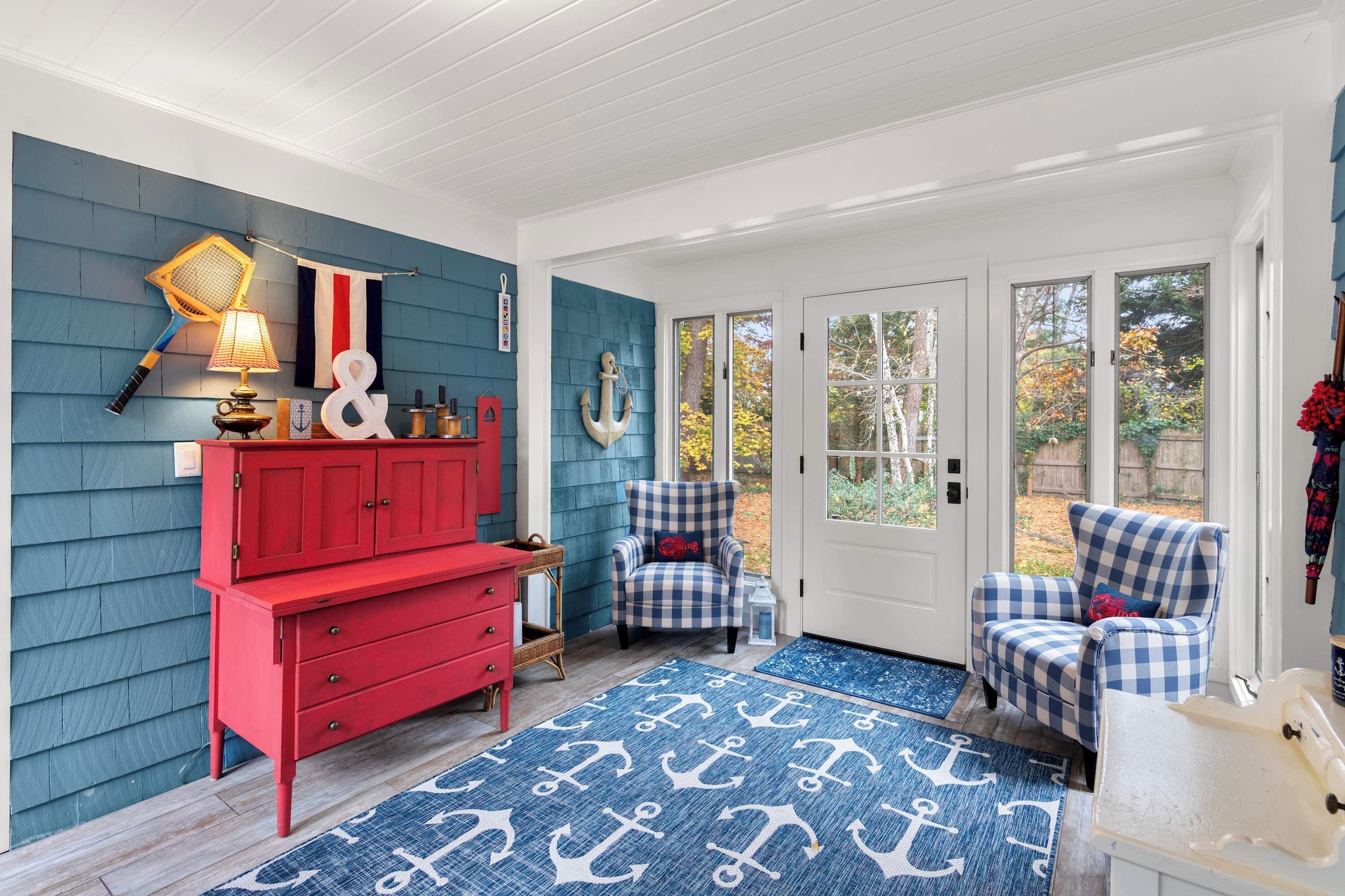 A room with a red dresser, two checkered armchairs, a blue and white patterned rug, and a door leading outside.