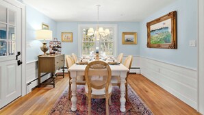A dining room with a white table set for six, a chandelier, and a large window with a view. - Game Room, Central AC, 4 mins to Craigville Beach! (Barnstable)