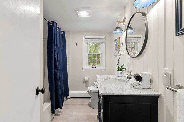 A bathroom with a white toilet, a sink with a marble countertop, a mirror, and a shower curtain. - Game Room, Central AC, 4 mins to Craigville Beach! (Barnstable)