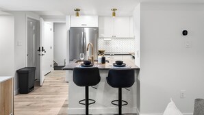 A modern kitchen with a stainless steel refrigerator, two black bar stools, and a herringbone tile backsplash.