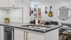 A modern kitchen with a granite countertop, stainless steel sink, and gold handles. There is a potted plant on the counter, and various kitchen items are visible.
