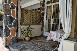 Ein überdachter Außenbereich mit Steinmauern, einem Blumentopf, einem Tisch mit einer Blumen Tischdecke und einem Fenster mit Vorhängen.