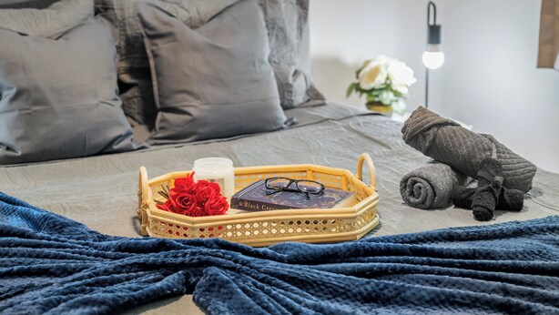 A neatly made bed with a blue blanket, a wicker tray with a book, a candle, and red roses.