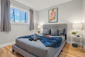 Ein Schlafzimmer mit einem Bett, Nachttischlampen, einem Fenster mit Vorhängen, einem Bild an der Wand und einem kleinen Tisch mit einer Pflanze.
