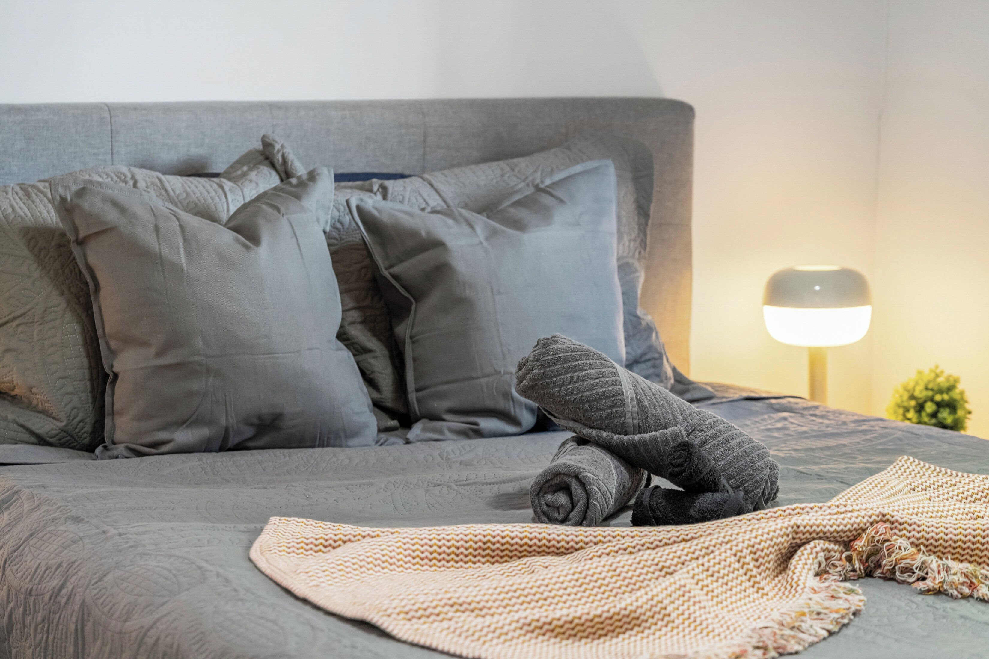 A neatly made bed with grey bedding and pillows, a rolled-up grey blanket, and a beige knitted throw.
