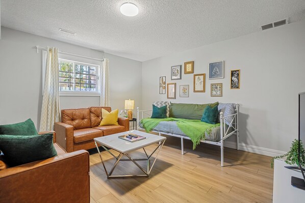 Ein Wohnzimmer mit braunem Ledersofa, einem weißen Couchtisch, einem weißen Sofa mit grüner Decke und einem Fenster mit Vorhängen.