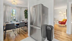 A modern kitchen with a stainless steel refrigerator, a dining area with a table and chairs, and a bright living space with a red chair and a lamp.